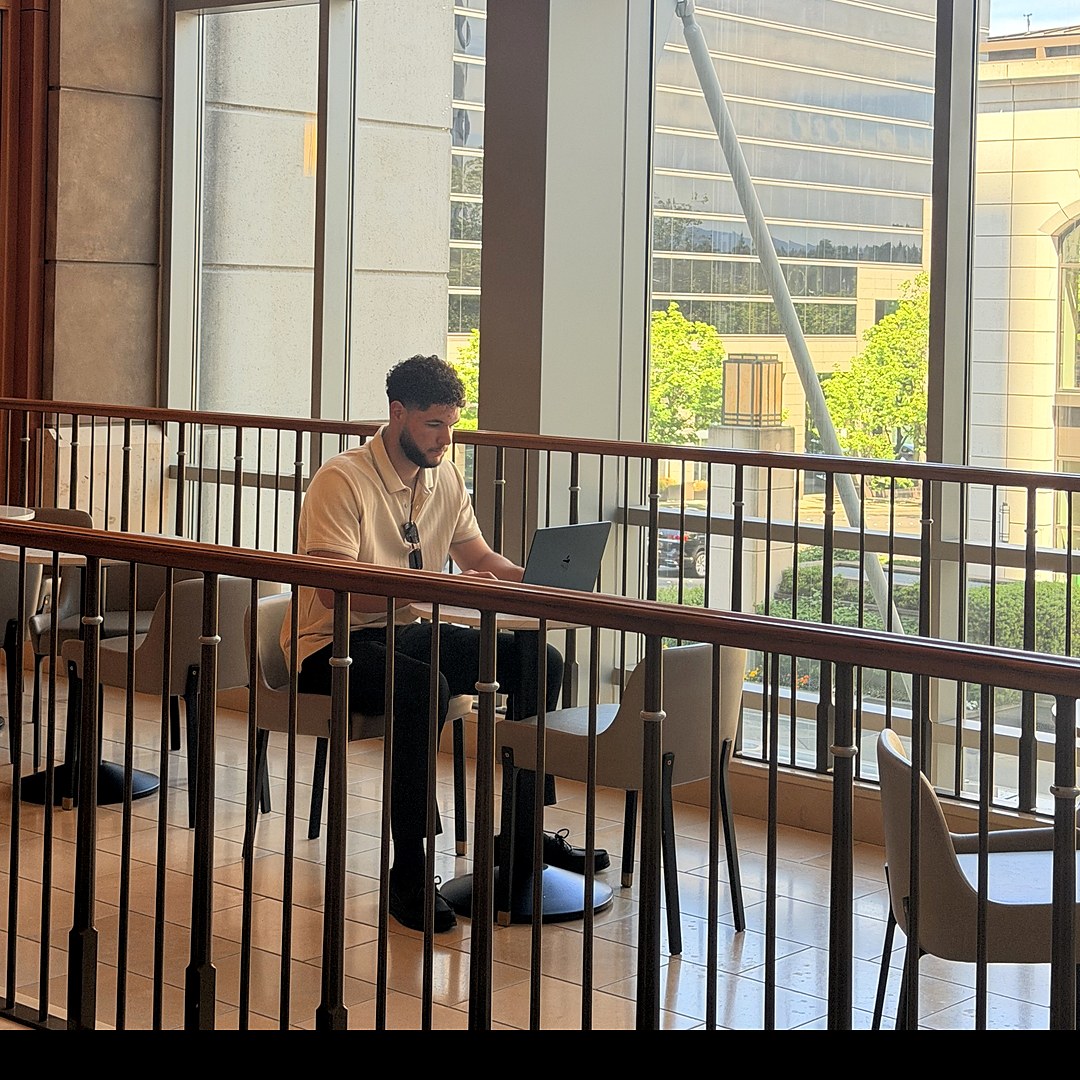 Joshua Daniels working on his laptop in a glass atrium overlooking a corporate campus